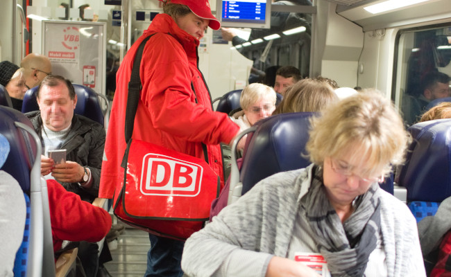 DB Promotorin in Roter Jacke und mit einer Roten Tasche wo das DB Logo drauf ist verteilt Schokolade im Zug