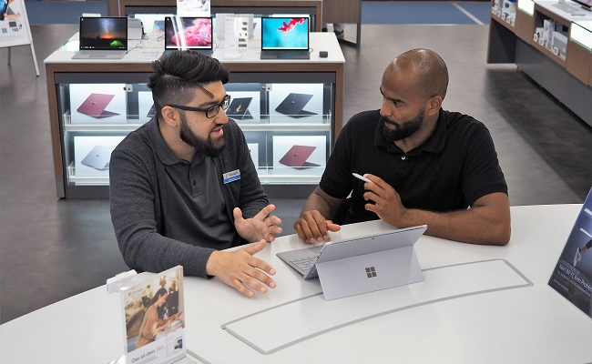Ein Mann in einem dunklen Hemd berät einen anderen Mann zu Surface Produkten von Microsoft an einem Tisch
