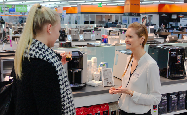 Zwei Frauen stehen in einem Laden vor diversen Kaffeemschinen und eine der beiden Frauen mit einem schwarz weißen Schal hält einen kleinen Becher mit Kaffee