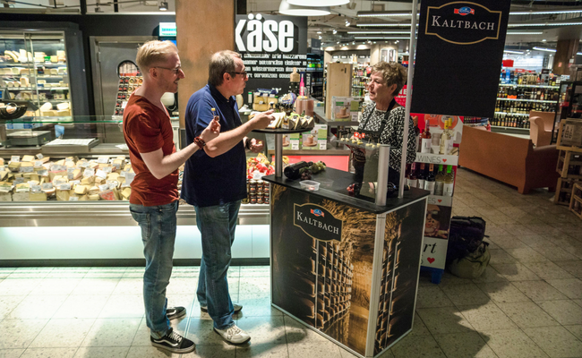 Zwei Männer stehen an einem Probierstand in einem Laden und hinter dem Stand steht eine Frau