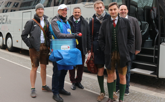 Ein Mann mit einer blauen Tasche steht neben einer Gruppe Männer welche kurze Hosen tragen vor einem Bus 