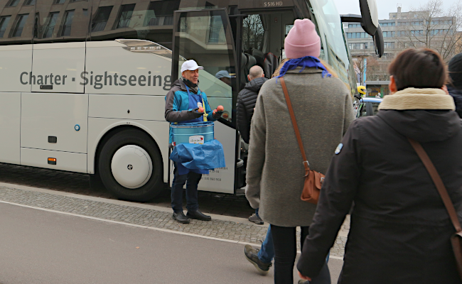 Ein Mann mit einer blauen Tasche steht an der Eingangstür einen Busses und verteilt Äpfel an die Leute die einsteigen