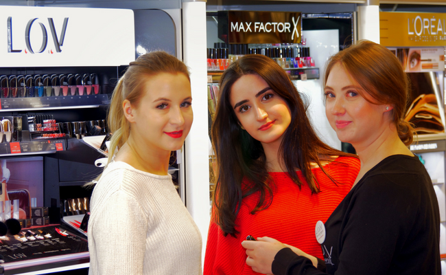 Drei Frauen stehen gruppiert vor einem LOV Regal in einem Laden und halten einen Lippenstift in der Hand