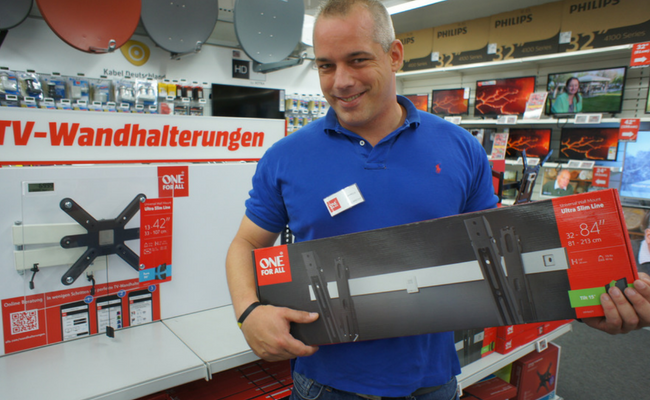 Ein Mann in einem blauen Polo hält eine verpackte TV-Wandhalterung in den Händen und lächelt