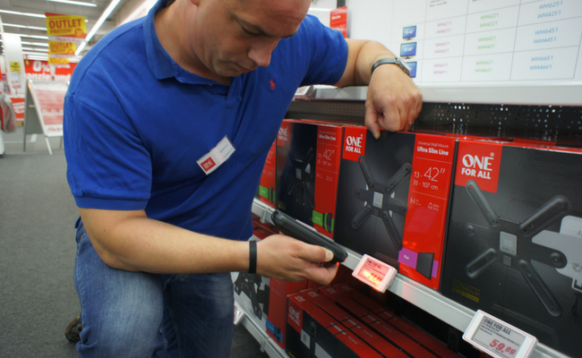 Ein Mann in einem blauen Polo scannt mit einem kleinen Gerät den Preis von einem Artikel in einem Laden