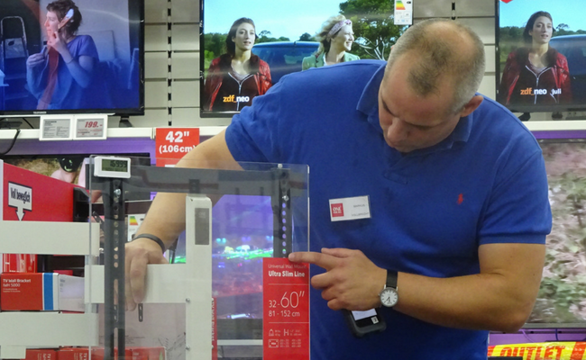 Ein Mann in einem blauen Polo steht in einem Laden und greift mit seiner Hand nach einem Produkt 