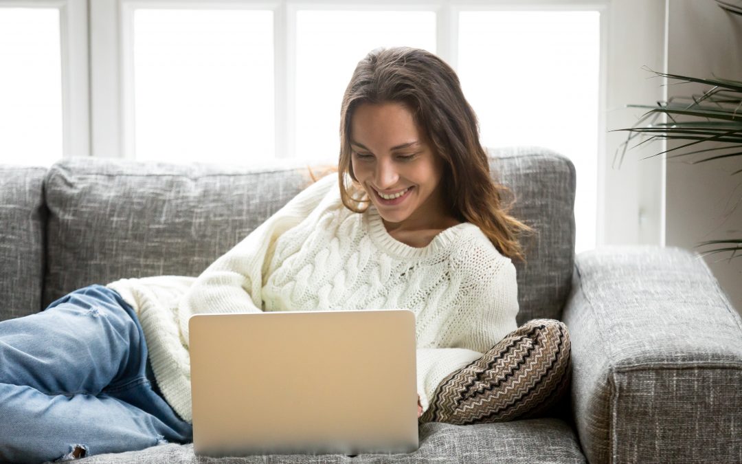Eine Frau mit einem weißen Strick-Pullover liegt auf dem Sofa mit dem Laptop vor sich und lächelt